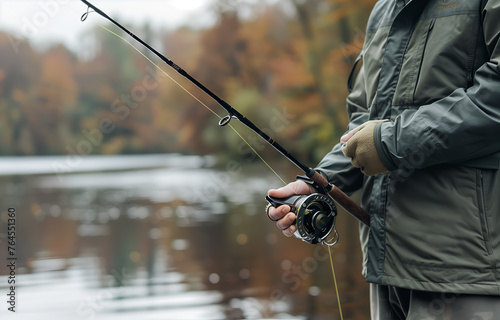 A closeup of the hands and fishing rod held by an outdoorsman, with blurred background showing river or lake in autumn season © digitalpochi