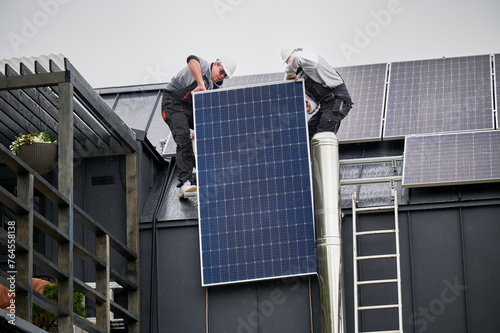 Men roofers installing solar panel system on roof of house. Technicians in helmets lifting up photovoltaic solar module with help of ropes outdoors. Concept of alternative and renewable energy.