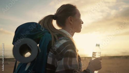 Woman tourist drinks water walks beach. Camper hiker female strolling along sea beach admiring nature at sunset. Carrying backpack with touristic equipment. Tourism vacations outdoors activity. photo