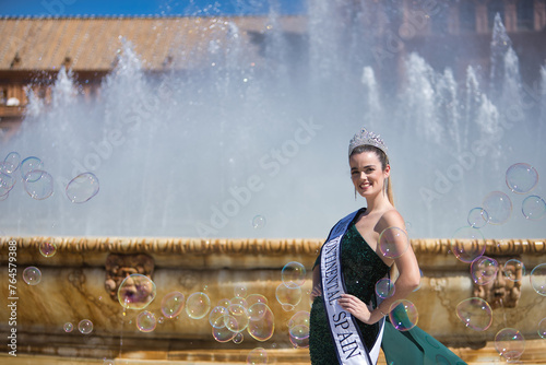 Young, pretty, blonde woman in a green party outfit with sequins, with a diamond crown and beauty pageant winner's sash, posing next to a fountain surrounded by soap bubbles.