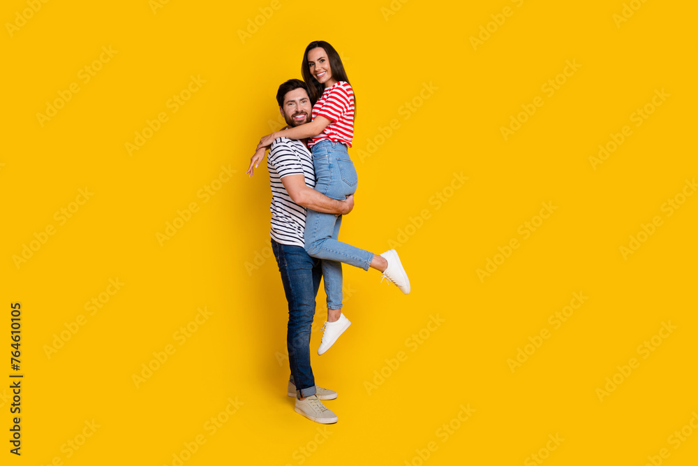 Full size photo of couple dressed striped t-shirt cuddle together man hold his girlfriend on arms isolated on yellow color background Full size photo of couple dressed striped t-shirt cuddle together man hold his girlfriend on arms isolated on yellow color background