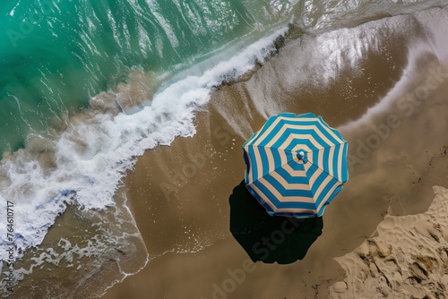 drone shot of bluestriped beach umbrella on sandy shore with turquoise waves nearby photo