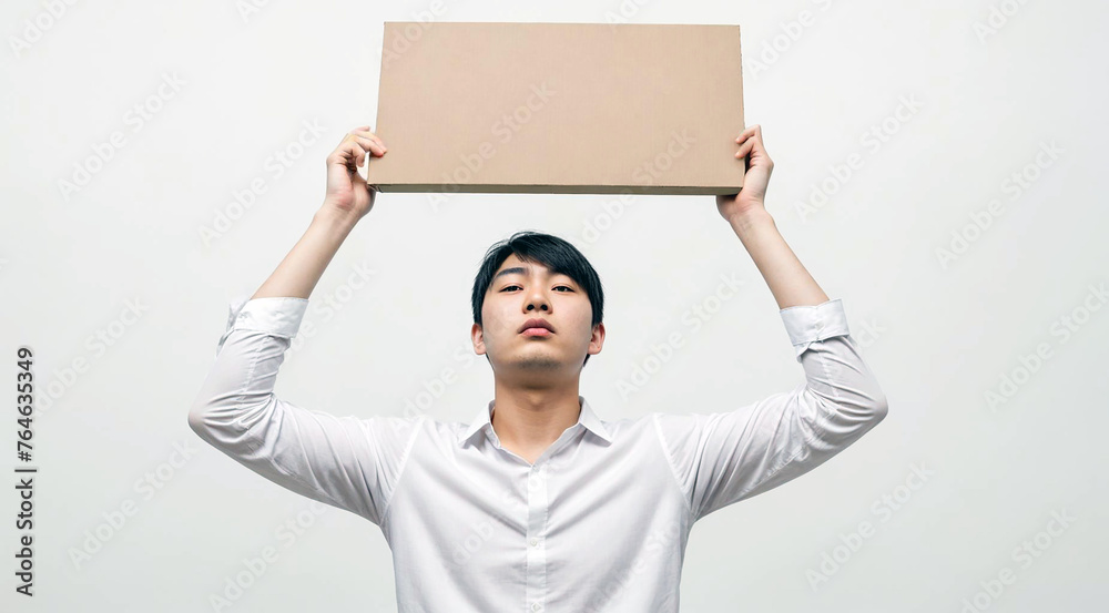 Young Adult Asian Man in White Shirt Holding a Big Empty Cardboard Sheet Above His Head. Image with Copyspace. Young Adult Asian Man in White Shirt Holding a Big Empty Cardboard Sheet Above His Head. Image with Copyspace.