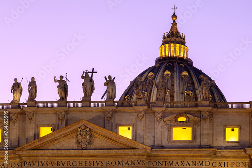 Details st at the rooftop of San Pietro Basilica (Saint Peter Cathedral) of Vatican City during dusk time, with statues and the illuminated dome of the cathedral.