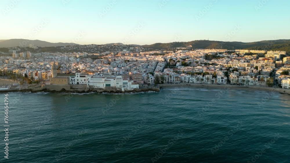 Panoramic aerial view of Sitges City. Drone tracking right. View of all region. Beautiful warm colours at sunset, reflecting on buildings. Travel destination in Spain. Panoramic aerial view of Sitges City. Drone tracking right. View of all region. Beautiful warm colours at sunset, reflecting on buildings. Travel destination in Spain.
