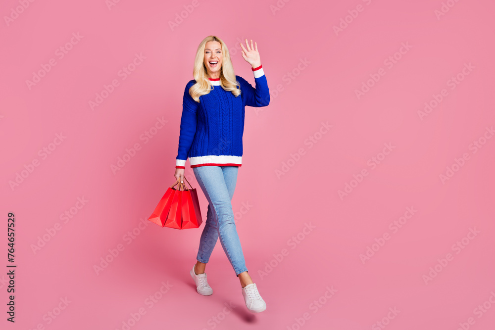 Full body portrait of pretty lady hold bags arm wave hi empty space wear blue sweater isolated on pink color background Full body portrait of pretty lady hold bags arm wave hi empty space wear blue sweater isolated on pink color background