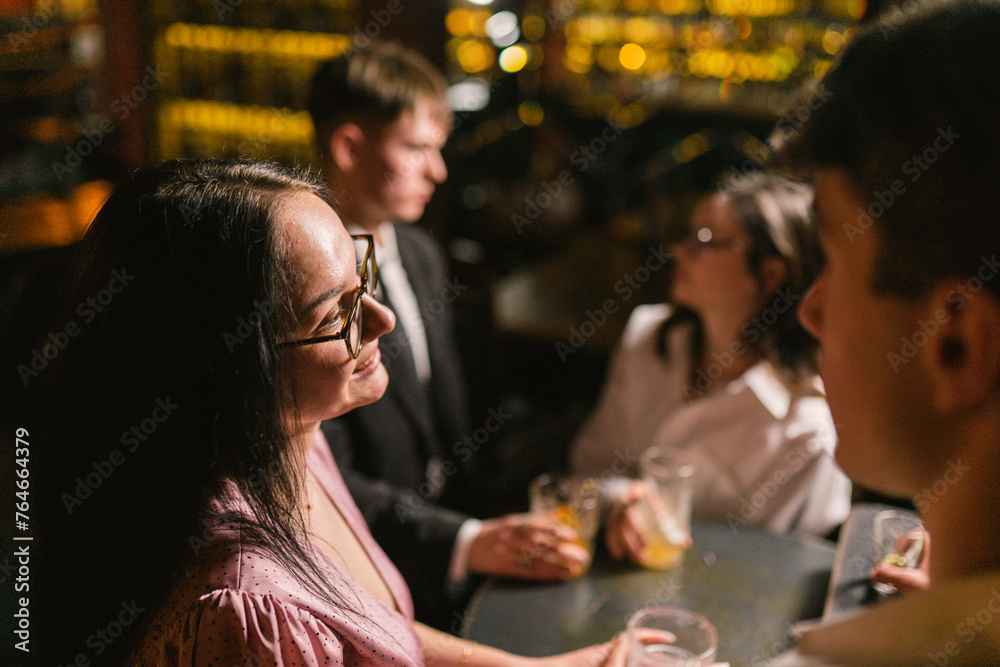 Brunette girl smiles at boyfriend against backdrop of dimmed bar with lamp lighting. Couples on double date in favorite pub