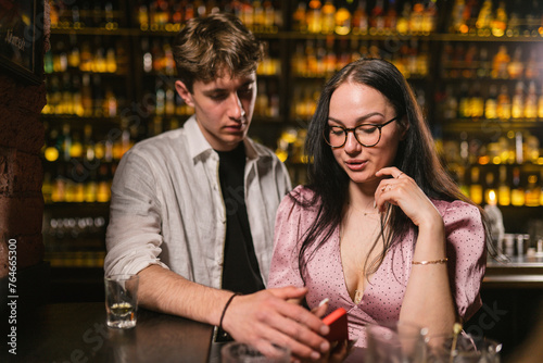 Young people sat at bar counter after fun party in trendy pub. Girl in elegant dress attracts attention of phone sitting in gloomy bar