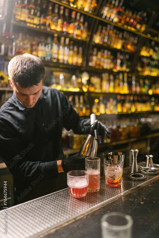 Professional barkeeper makes delicious cocktails for visitors of elite nightclub. Young barman in uniform makes efforts to earn more money