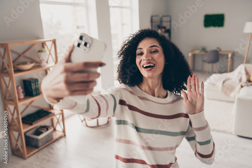 Photo of sweet excited lady dressed striped pullover recording video modern device waving arm hi indoors apartment room