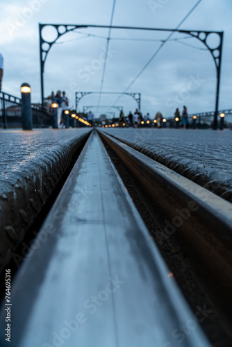 Porto metro train or tram track in infinite perspective at ground level of the Dom Luis bridge at sunset, under a cloudy sky and silhouettes of tourists walking in the background.