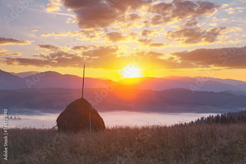 Bright colorful sunrise over foggy mountain valley. Panoramic landscape with a beautiful sky with clouds, rising over Carpathian mountains. Haystack on grassland hill in the foreground.