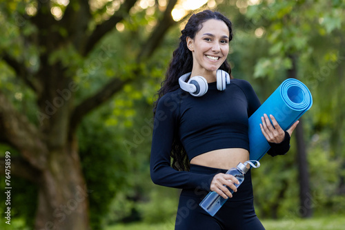 Fototapeta Naklejka Na Ścianę i Meble -  A smiling, active woman holding a yoga mat and water bottle while wearing headphones in a peaceful park setting.