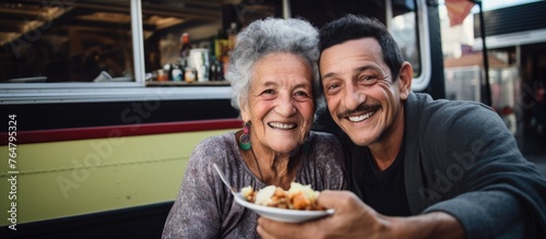 An elderly man and woman smiling while holding a plate of food in front of a bus