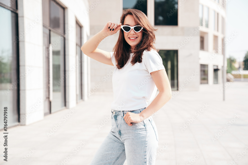 Young beautiful smiling hipster woman in trendy summer white t-shirt and jeans clothes. Sexy carefree model posing on the street background at sunset. Positive model outdoors. In sunglasses Young beautiful smiling hipster woman in trendy summer white t-shirt and jeans clothes. Sexy carefree model posing on the street background at sunset. Positive model outdoors. In sunglasses