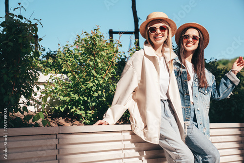 Two young beautiful smiling hipster female in trendy summer clothes. Carefree women posing in the street. Positive models having fun outdoors at sunny day. Cheerful and happy. In hat and sunglasses