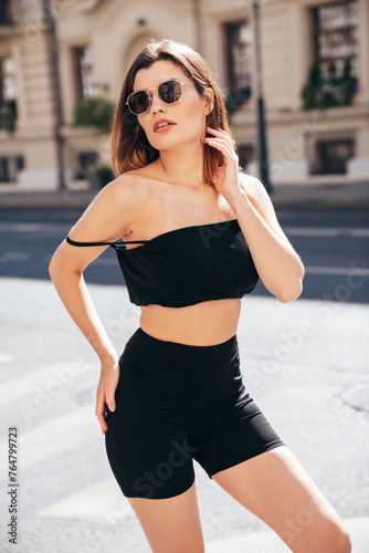 Young beautiful confident female in trendy summer black cycling shorts and tank top clothes. Sexy carefree woman posing in the street. Positive model. Cheerful and happy. In sunglasses