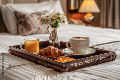 Bandeja de madera con la inscripción happy mother day conteniendo desayuno con taza de café, zumo de naranja y cruasan y flores, sobre una cama y fondo de habitación desenfocado