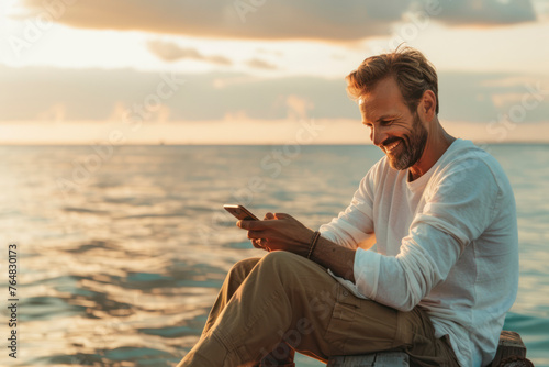 Happy man text messaging through smart phone while sitting on pier by sky and sea.