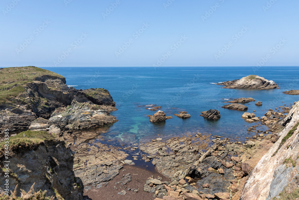 A scenic coastline with rocky formations, blue waters, and distant mountains under a clear sky, exhibiting natural beauty