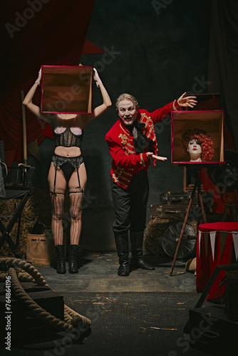 Tricks. Magician in red coat showing tricks with headless female assistant holding boxes over dark retro circus backstage background. Concept of circus, theater, performance, show, retro and vintage