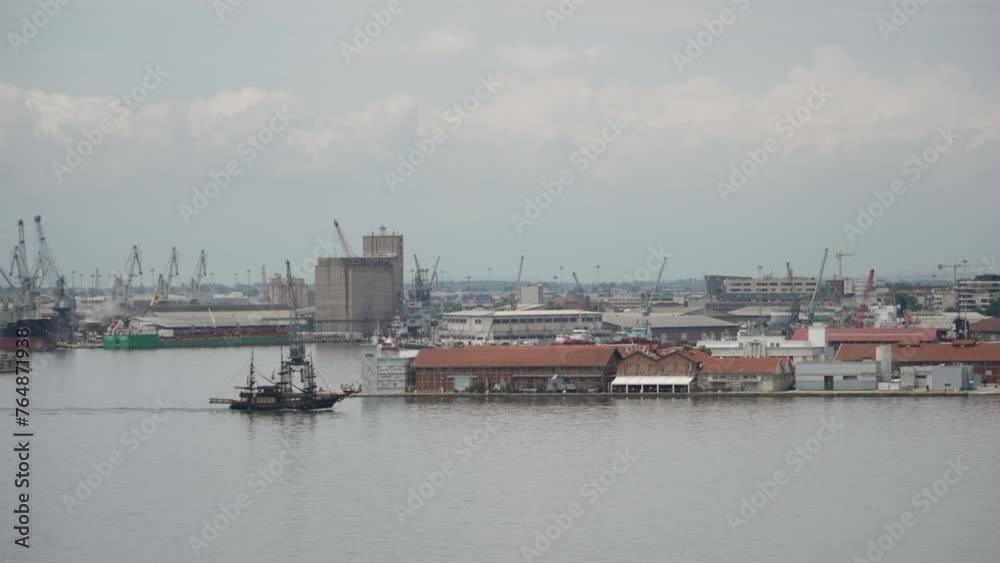 Tourist old pirate ship made of dark wood sails near the cargo port. Industrial landscape of sea trading bay, wide shot. Marine transport links in the city on sunny summer day. Tourist old pirate ship made of dark wood sails near the cargo port. Industrial landscape of sea trading bay, wide shot. Marine transport links in the city on sunny summer day.