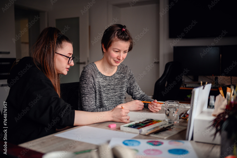 In a cozy, cluttered workspace, two women share a light-hearted moment over their art projects, surrounded by creative tools