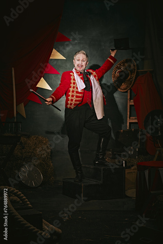 Emotional man with pale face makeup, in red stage costume and hat making performance over dark retro circus backstage background. Concept of circus, theater, performance, show, retro and vintage photo