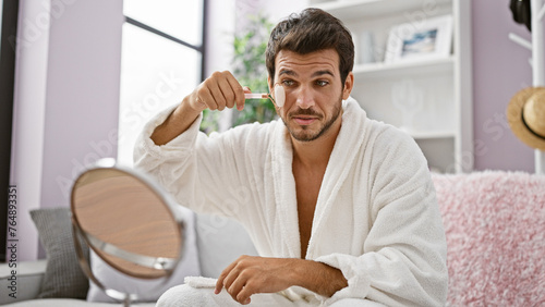 A handsome man in a white bathrobe grooming with a jade roller while sitting at home.