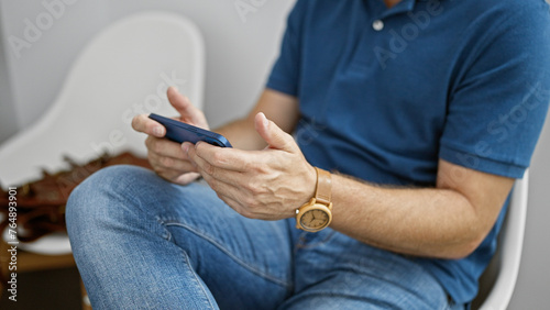 Mature hispanic man using smartphone in a modern indoor room  wearing casual clothing and a wristwatch.