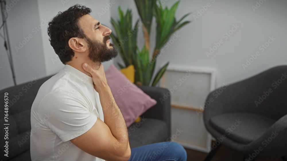 A bearded man in a white t-shirt experiencing neck pain in a modern living room A bearded man in a white t-shirt experiencing neck pain in a modern living room