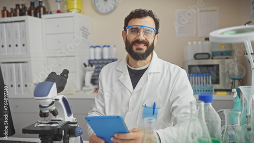 Smiling bearded man in lab coat and safety glasses holding a tablet inside a laboratory filled with equipment