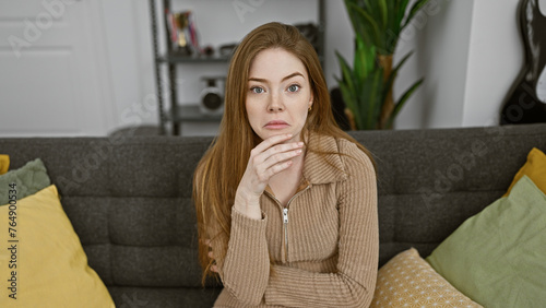 Caucasian woman with blonde hair wearing a sweater sitting thoughtfully on a couch indoors, portraying a comforting home scene.