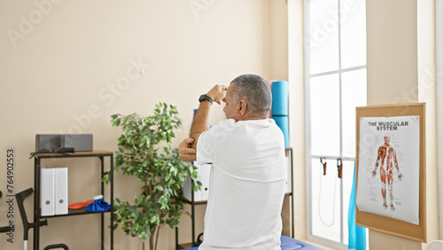A mature hispanic man stretching his arm in a rehab clinic room, illustrating health and therapy.