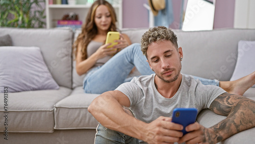 A tattooed man and a woman engage with their smartphones on a gray couch in a modern living room.