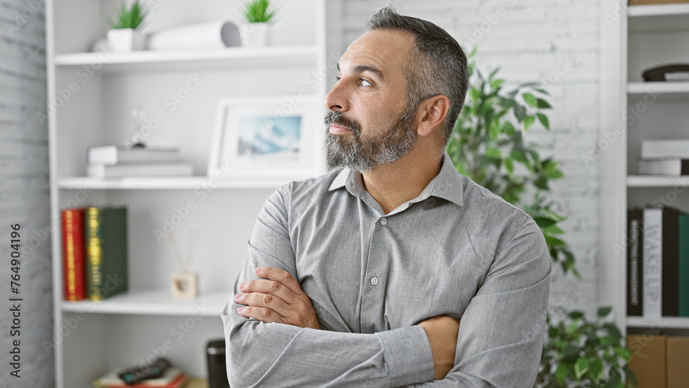 Fototapeta premium Hispanic senior man with beard and grey hair standing arms crossed in a modern office interior.