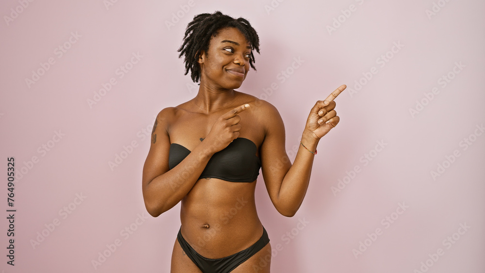 Confident young woman with dreadlocks pointing to the side against a pink wall in casual black attire. - obrazy, fototapety, plakaty 