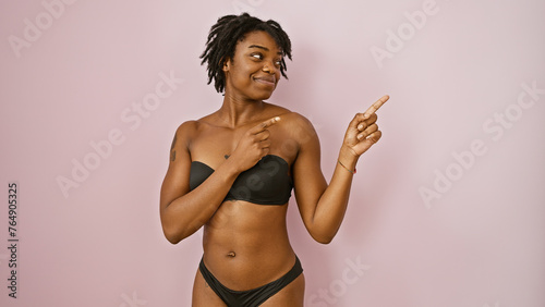Confident young woman with dreadlocks pointing to the side against a pink wall in casual black attire.