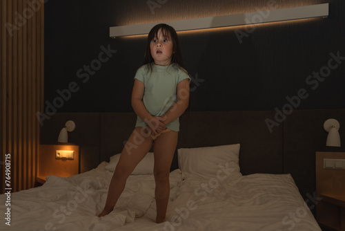 A little girl child with a frightened face stands on the bed in her parents’ bedroom. photo