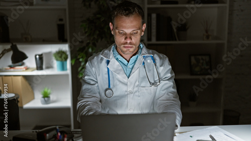 Hispanic male doctor in lab coat working on laptop at nighttime in clinic office, evoking dedication. photo