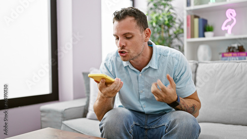 Angry young hispanic man with a beard in casual attire using a smartphone in a modern living room.