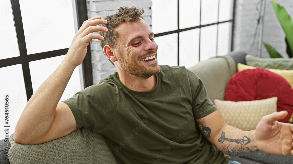 Laughing young adult hispanic man with beard and tattoo comfortable in living room home interior Laughing young adult hispanic man with beard and tattoo comfortable in living room home interior
