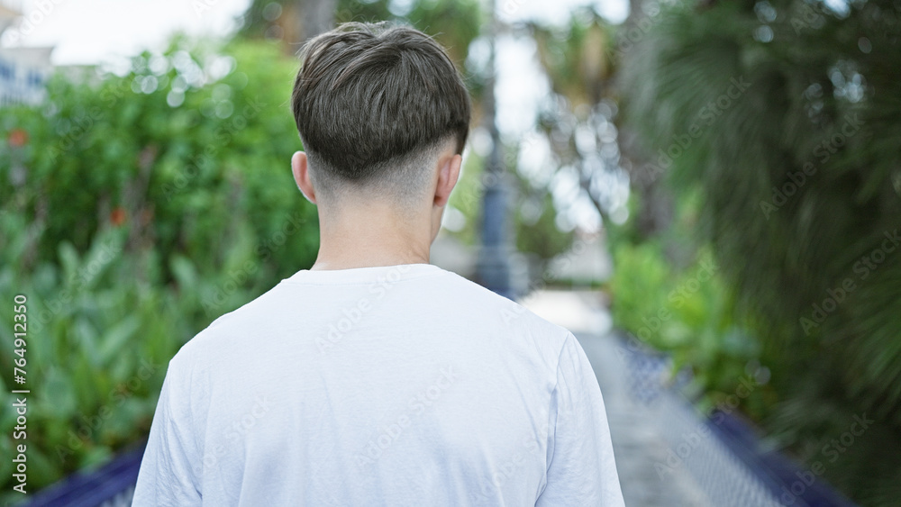 Rear view of a young caucasian man in a white shirt standing outdoors amidst lush greenery on a sunny day. Rear view of a young caucasian man in a white shirt standing outdoors amidst lush greenery on a sunny day.