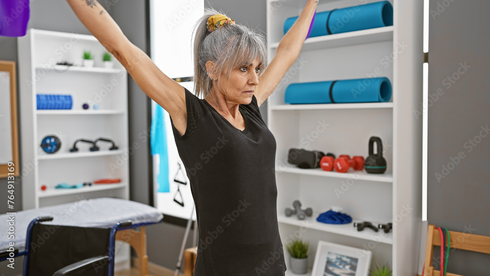 A mature hispanic woman exercising with outstretched arms in a rehab clinic's gym, indoors. A mature hispanic woman exercising with outstretched arms in a rehab clinic's gym, indoors.