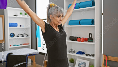 A mature hispanic woman exercising with outstretched arms in a rehab clinic's gym, indoors. photo
