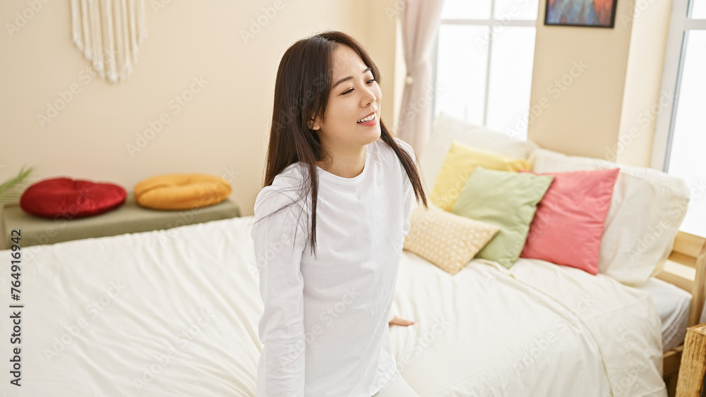 A smiling young asian woman sits in a well-lit, cozy bedroom with colorful pillows and modern decor, exuding casual comfort and style. A smiling young asian woman sits in a well-lit, cozy bedroom with colorful pillows and modern decor, exuding casual comfort and style.