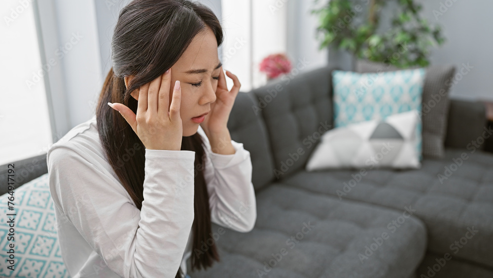 A stressed chinese woman with a headache at home, sitting on a couch in a modern living room. A stressed chinese woman with a headache at home, sitting on a couch in a modern living room.