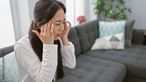 A stressed chinese woman with a headache at home, sitting on a couch in a modern living room.