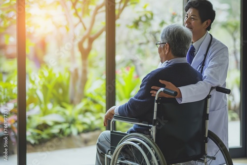 Middle-aged Asian doctor pushes his old gray-haired patient in a wheelchair indoors..