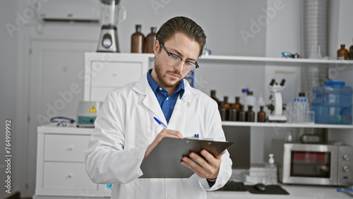 Young hispanic man scientist writing report standing at laboratory